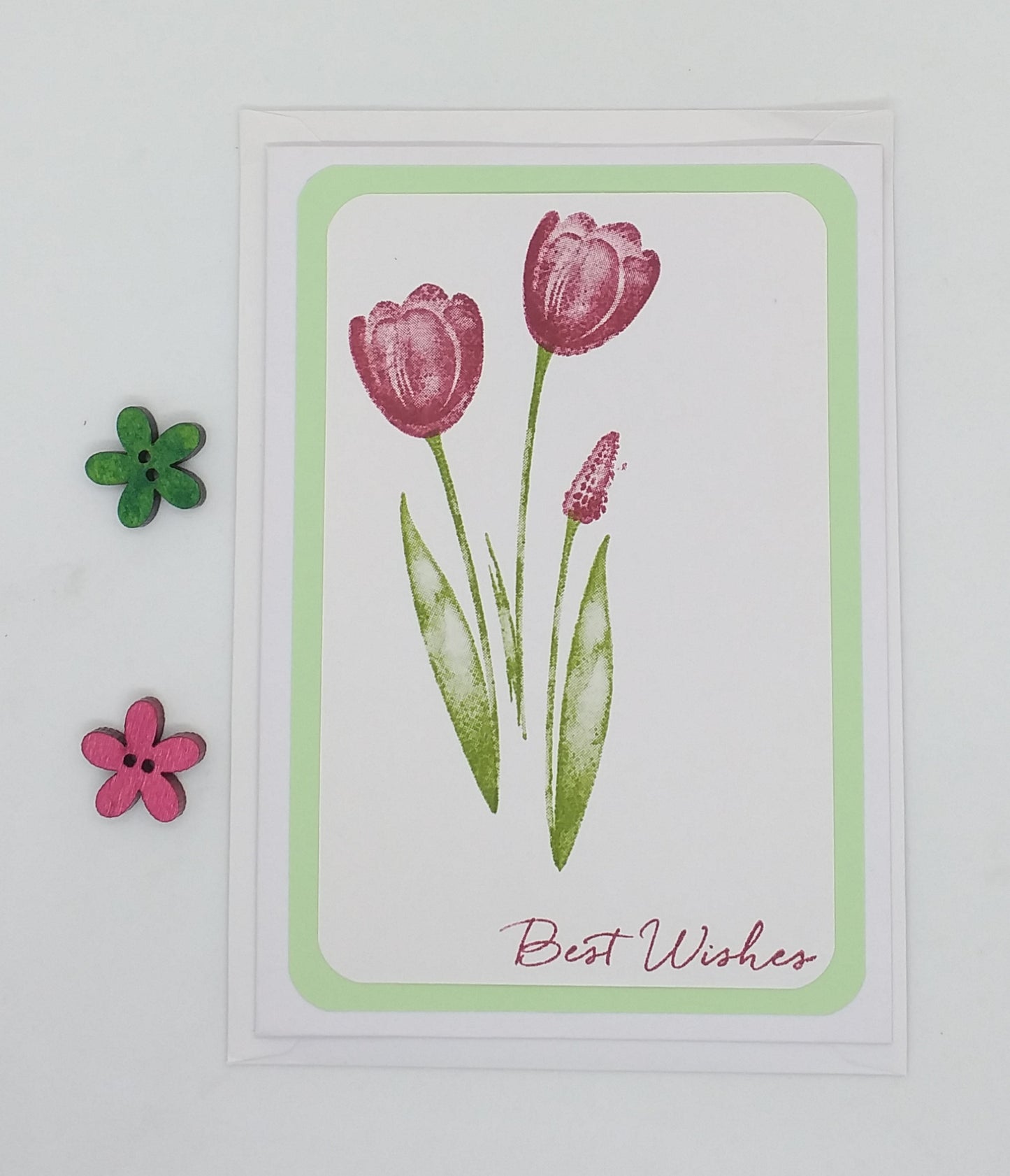 A greeting card with an envelope, featuring a hand-stamped design of tulips in shades of pink and green on a white background, with the words 'Best Wishes' written at the bottom.