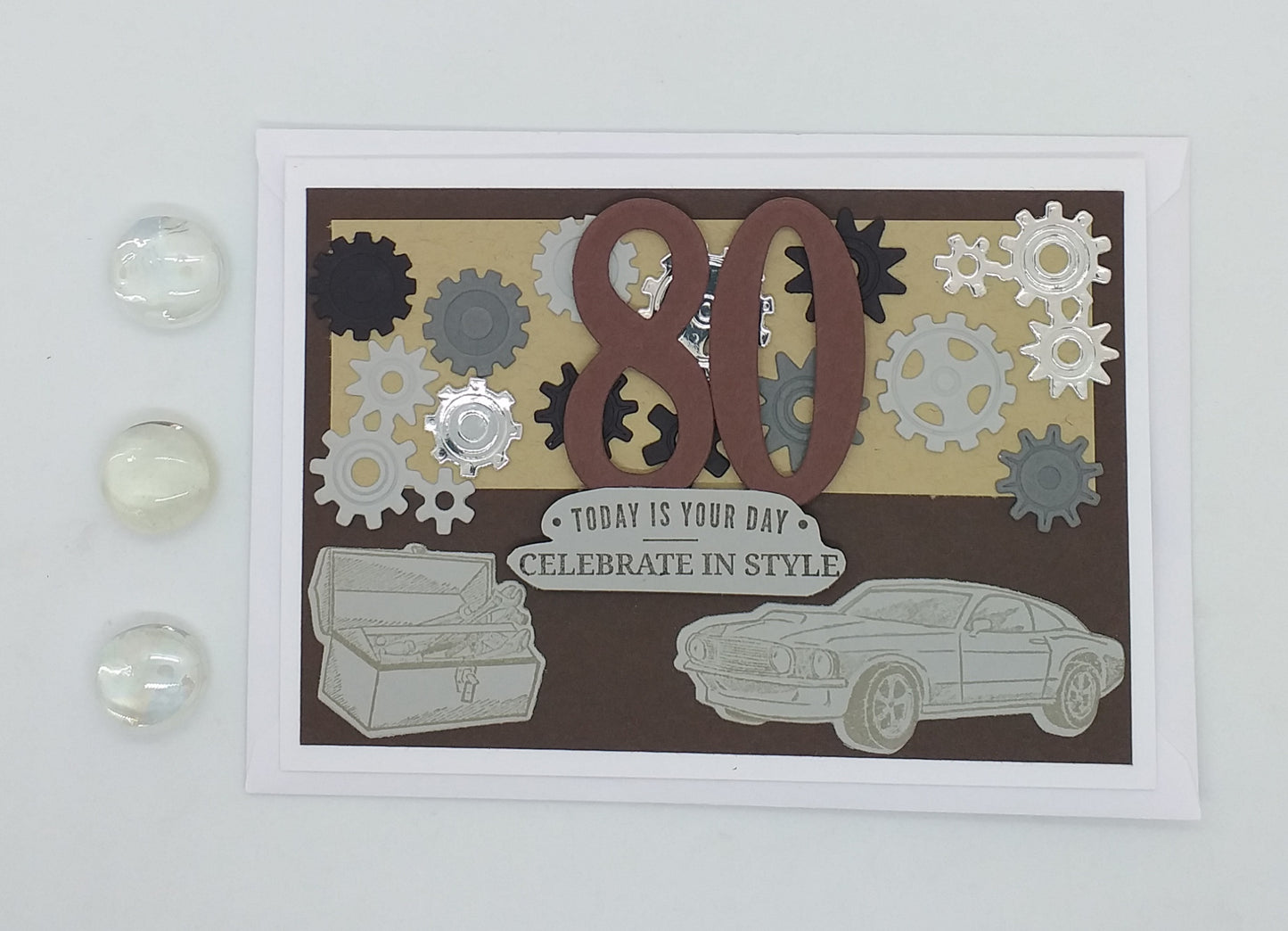 A greeting card with a vintage car and cogs design, featuring text for an 80th birthday celebration, accompanied by an envelope and two decorative stones.
