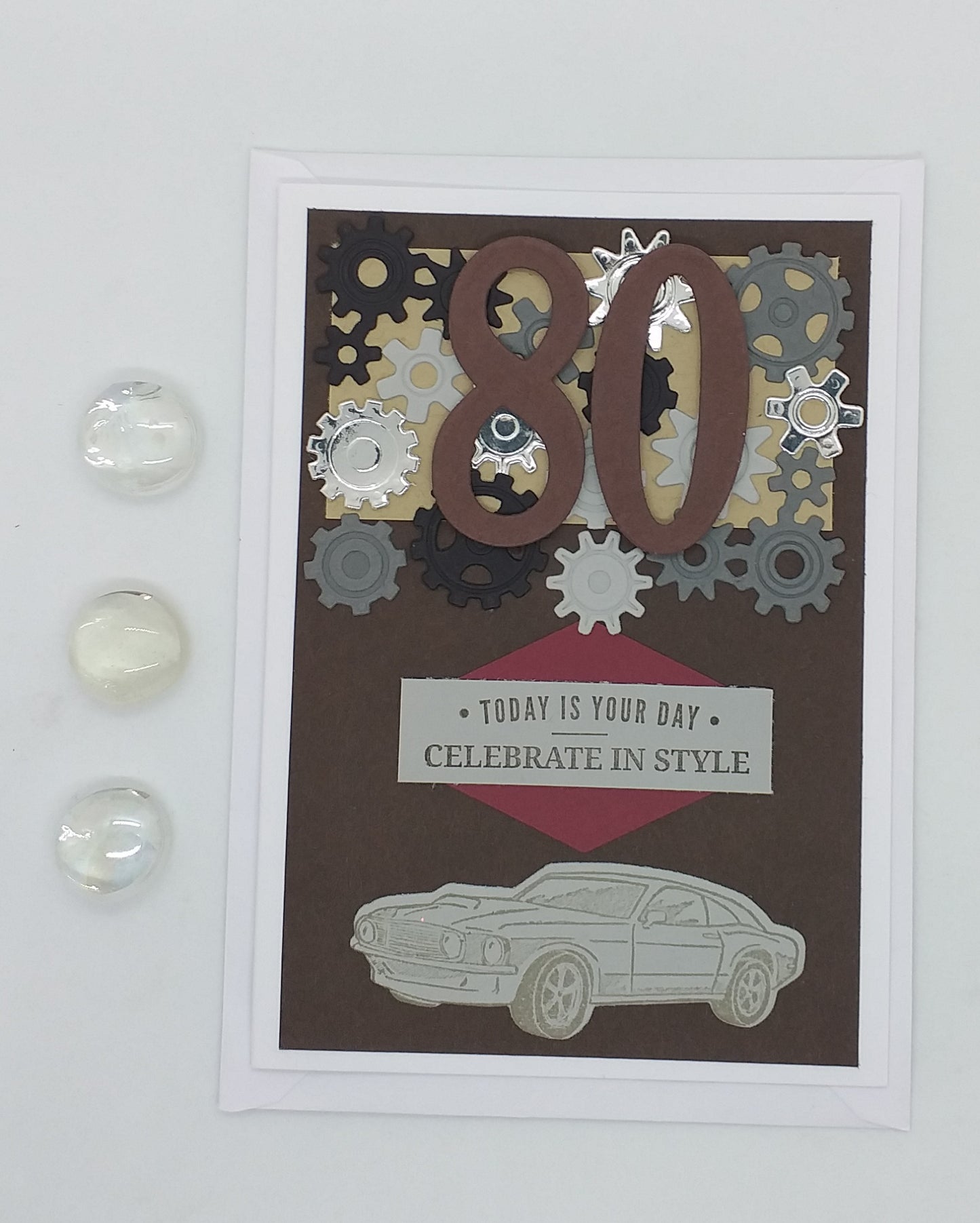 A greeting card with a birthday theme, featuring a layered cog design, the number 80, and the text 'Today is for style, celebrate in style'. There is an envelope to the side and a small, clear, spherical ornament on the front.
