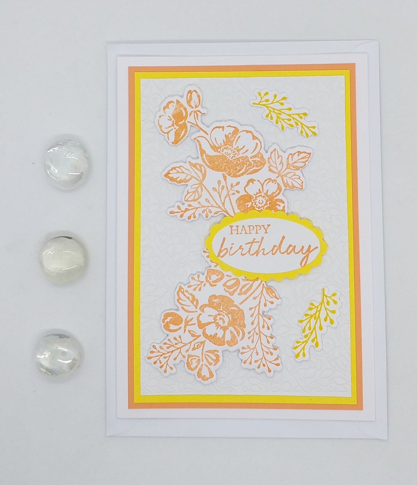 A cheerful floral birthday card with 'Happy Birthday' text in the center, surrounded by orange and yellow flowers, presented with two clear marbles on the side.