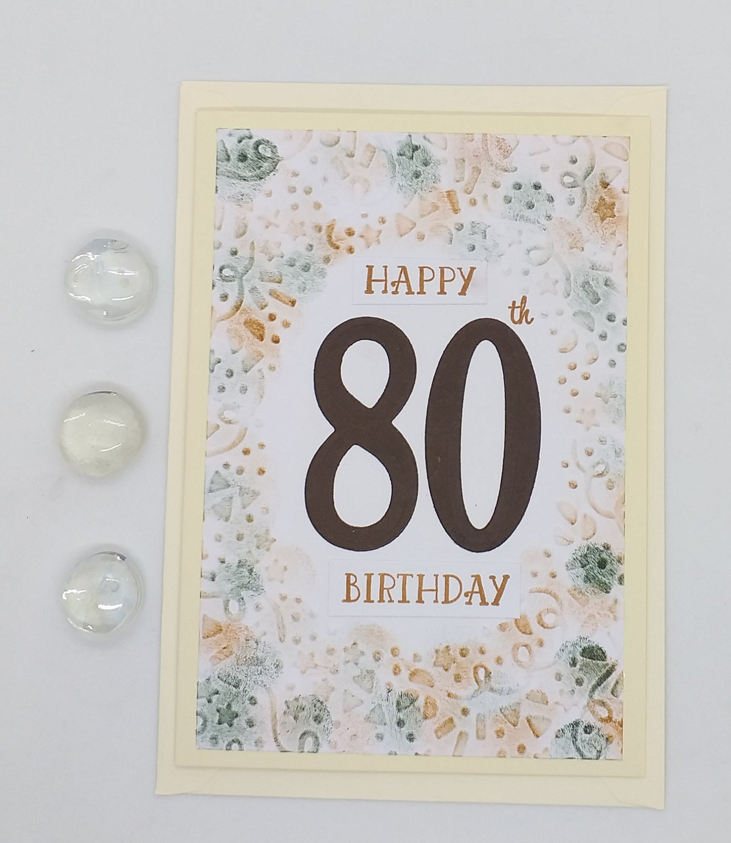 An A6 greeting card with 'Happy 80th Birthday' text embossed on it, along with some decorative elements, presented with an envelope and two small crystal-like embellishments.