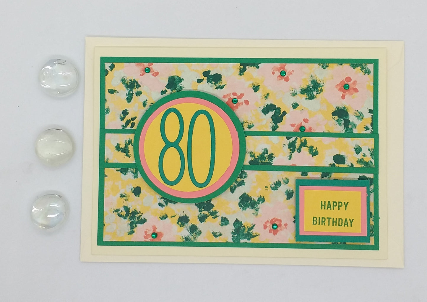 A colorful floral birthday card with the number 80 and the text 'Happy Birthday' displayed prominently on the front. The card is accompanied by two clear glass embellishments.