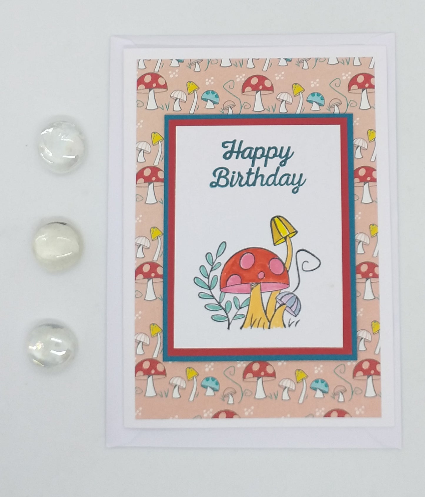 A greeting card with 'Happy Birthday' text, decorated with colorful mushroom illustrations and a small, clear ornament on the front, accompanied by a matching envelope.