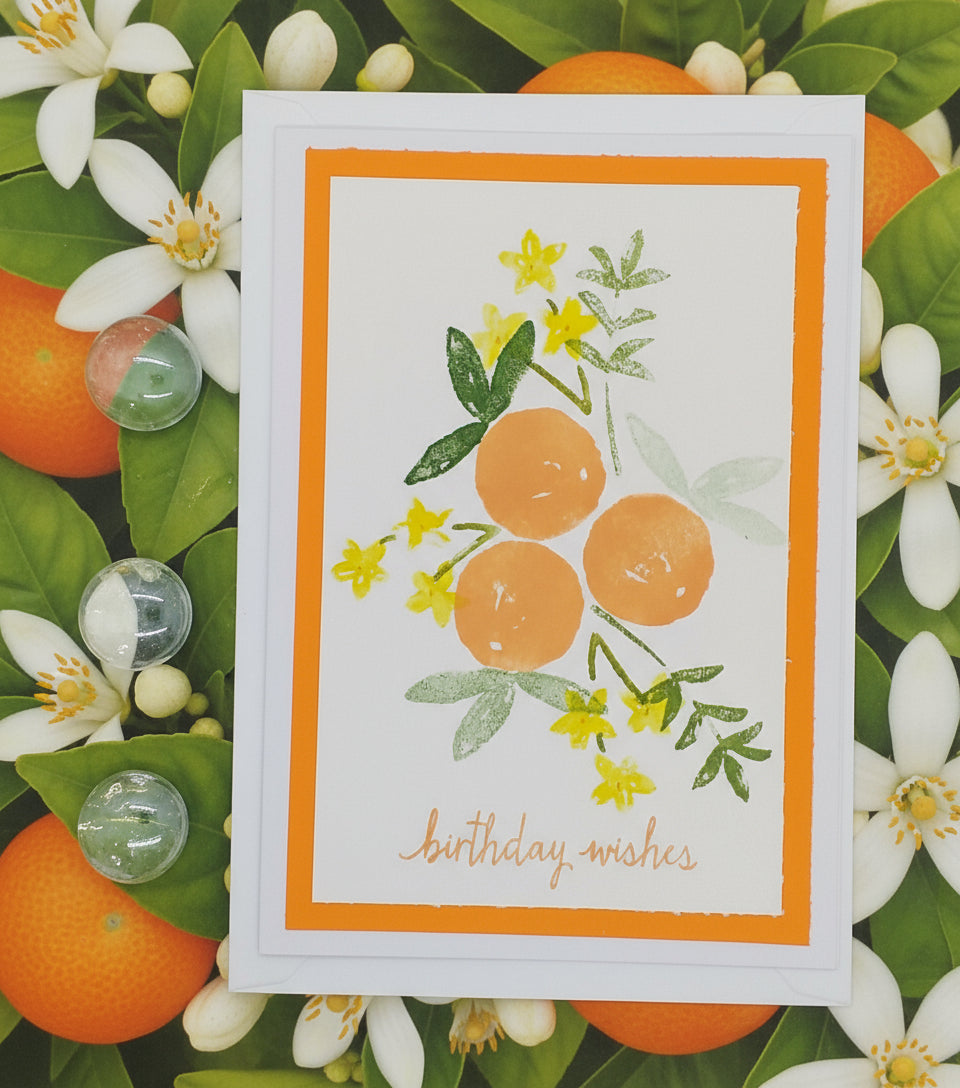Hand stamped and layered a6 card with an orange background, featuring illustrations of orange blossoms and green leaves, and the text 'Happy Birthdays'.