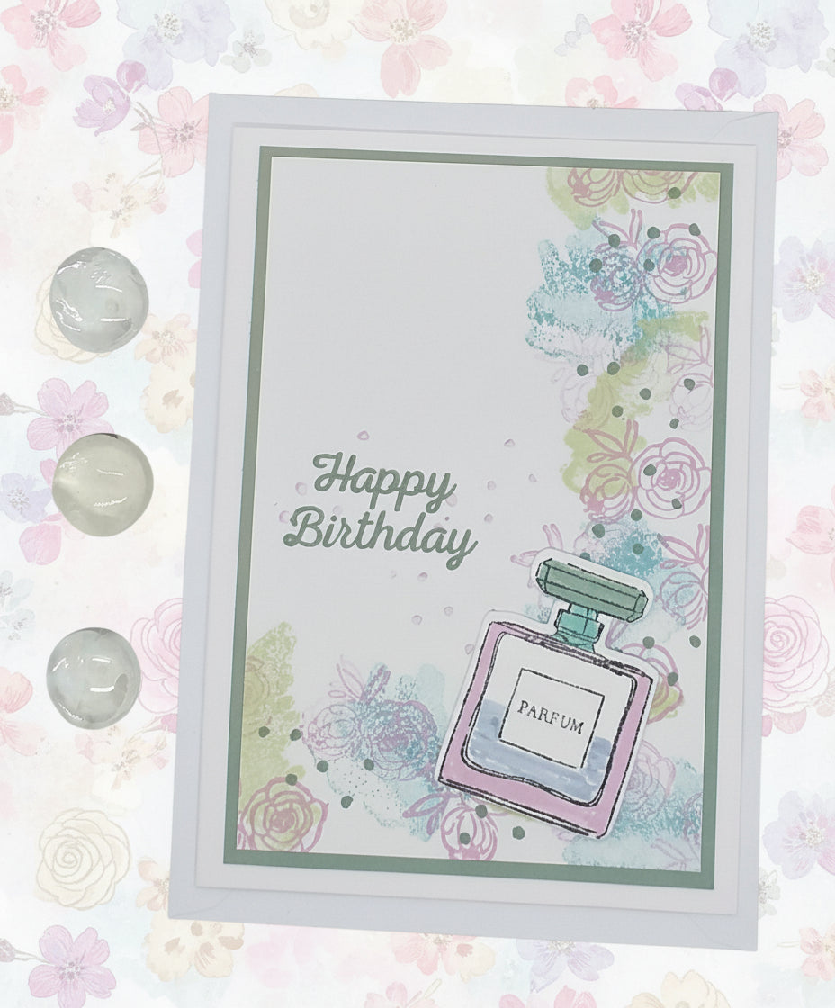 A greeting card with a stamped and layered design of a perfume bottle, featuring the words 'Happy Birthday' and decorated with colorful flowers and a crystal embellishment.