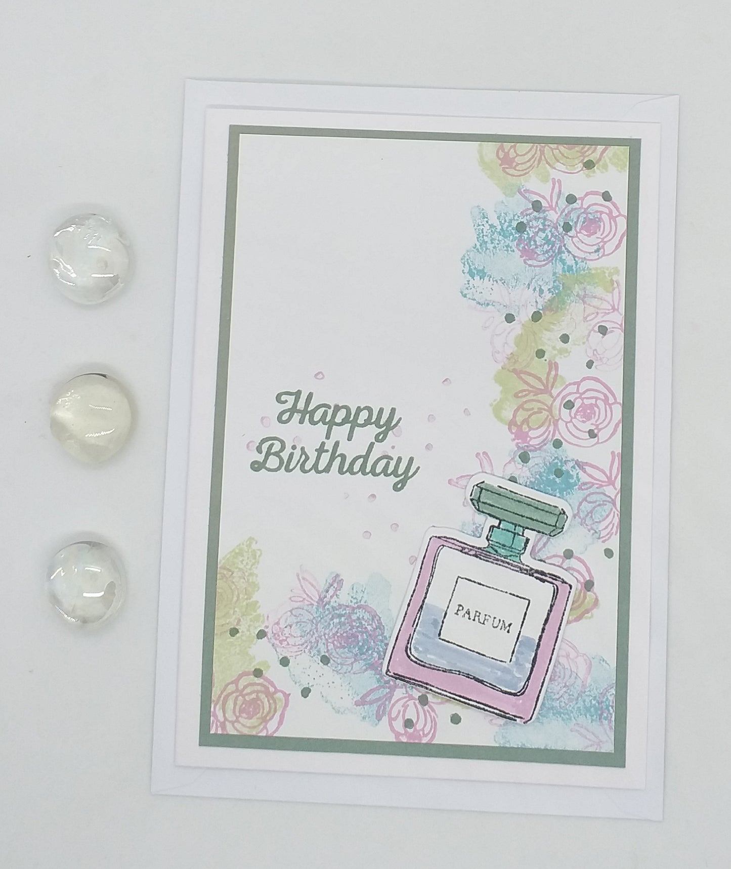 A greeting card with a stamped and layered design of a perfume bottle, featuring the words 'Happy Birthday' and decorated with colorful flowers and a crystal embellishment.