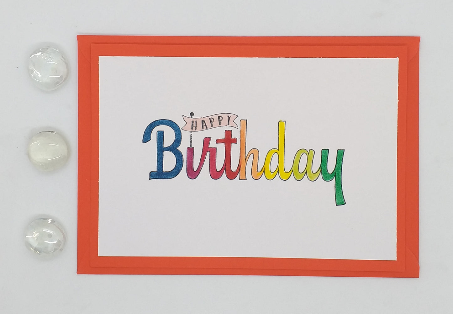A vibrant birthday card with a colorful 'Happy Birthday' greeting in a Display font, presented with a red envelope and two decorative elements on the side.