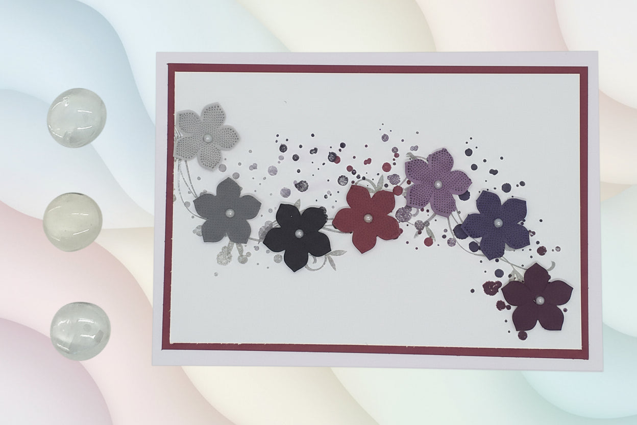 A greeting card with a floral design, featuring a row of curved flowers in shades of purple, gray, and red, with dainty pearls in the centre of each flower. The card has a blank inside for a personalized message and comes with an envelope.