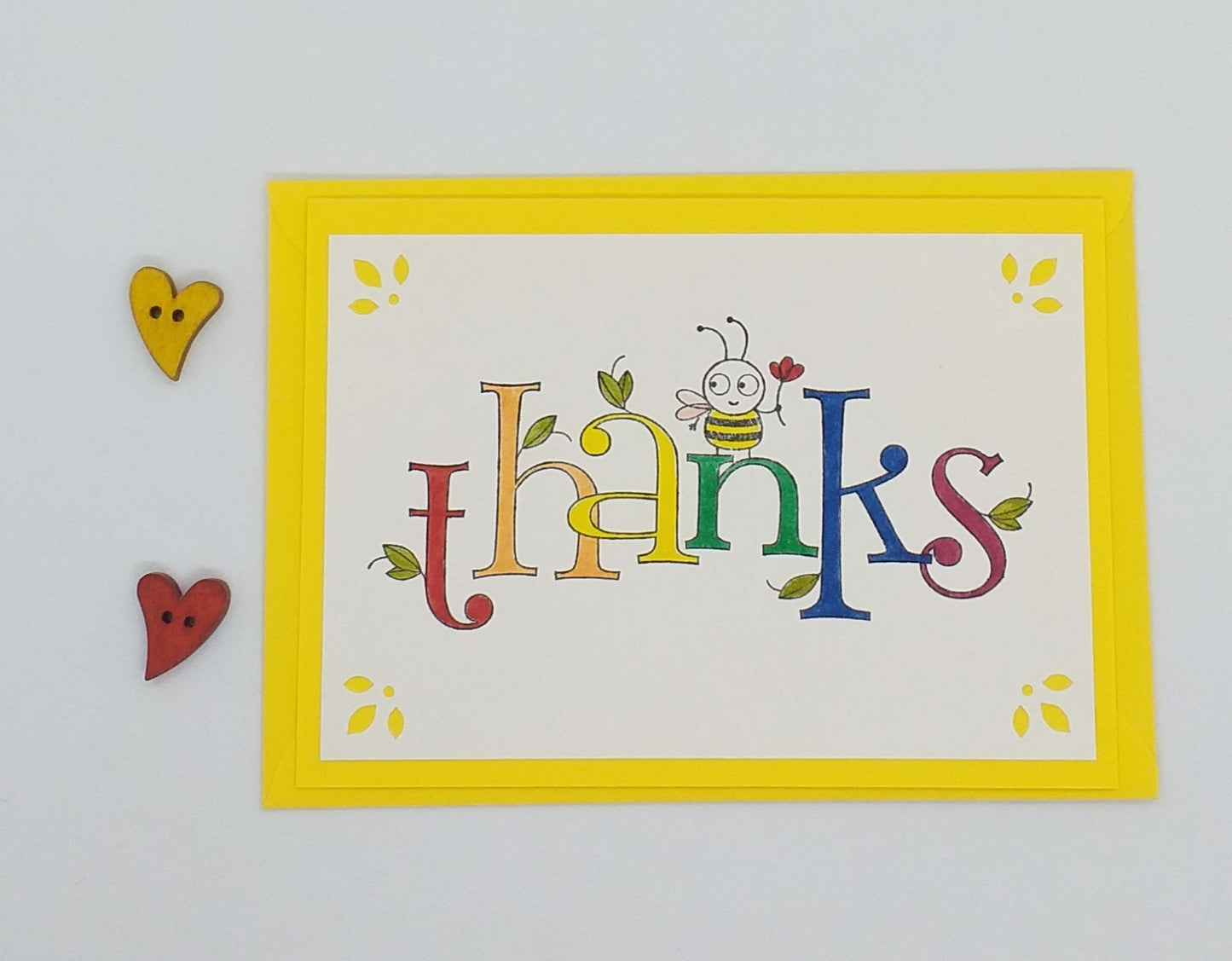 A colorful a6 card with the word 'thanks' written in bold letters, featuring a happy bee illustration, accompanied by a bright yellow envelope, all laid out on a white surface.