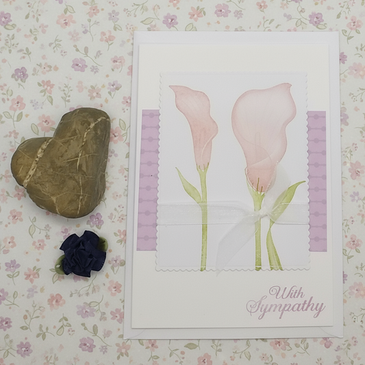 Sympathy card with pink flowers and 'With Sympathy' text on a floral background, accompanied by a heart-shaped stone and black rose.