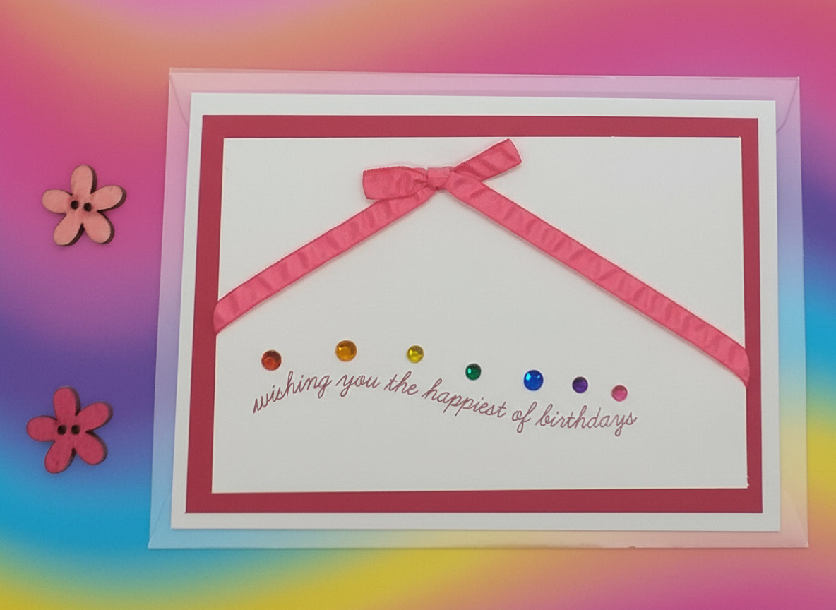 Hand-stamped birthday card with pink and pinker shades, decorated with ribbons and gems, and a written message that reads 'Wishing you the happiest of birthdays'.