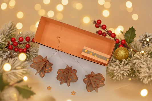 Three hanging decorative ornaments shaped like brown leaves with orange polka dots are displayed below an open orange box, with two small orange star-shaped embellishments on a white textured background.