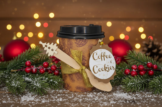A brown paper coffee cup with a black lid, decorated with a gold glitter ribbon, a wooden Christmas tree ornament, and a round tag that reads 'Coffee & Cookies'.