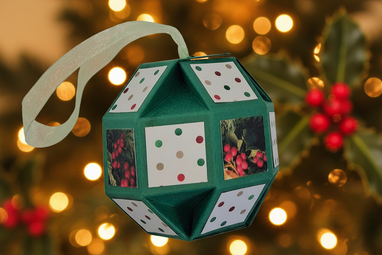 A green faceted geometric Christmas ornament with white panels featuring colorful dots and berry prints, hanging from a sheer pale green ribbon.