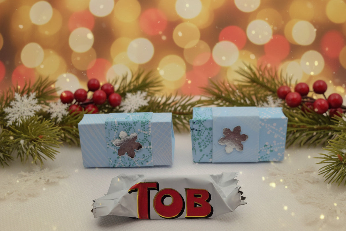 Two small light blue patterned boxes with flower-shaped cutouts are shown behind a wrapped candy bar featuring the letters 'TOB' in red with a gold outline on a white wrapper, all on a white surface.