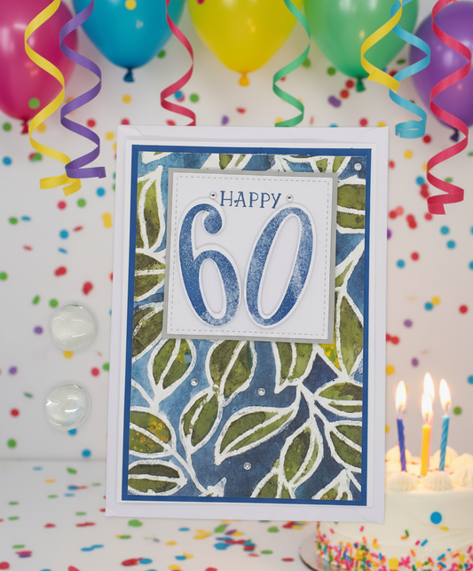 A birthday card with 'HAPPY 60' written on it, set against a patterned background, accompanied by two transparent envelopes.
