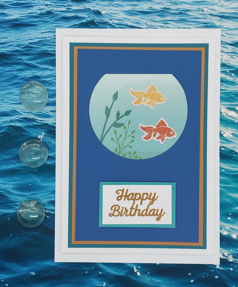 A greeting card with a hand-stamped image of two goldfish in a bowl, with 'Happy Birthday' text at the bottom. The card is displayed alongside its envelope, which is empty and plain.
