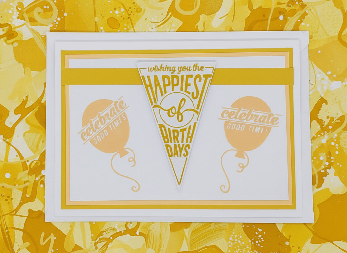 A6-sized birthday card with 'Happy Birthday' text in a yellow font, accompanied by an illustration of two balloons, one in a lighter shade and the other in a darker shade, placed on a white background. The card also features a small, clear crystal decoration.